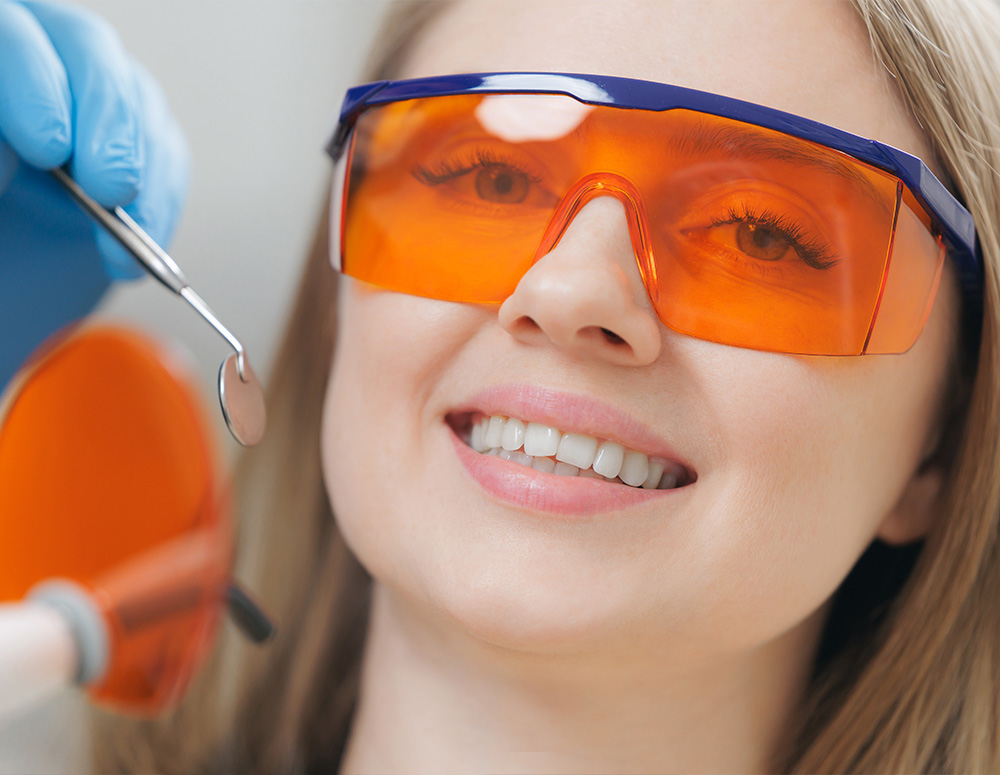 A woman wearing safety goggles and holding a pair of dental tools, smiling at the camera.